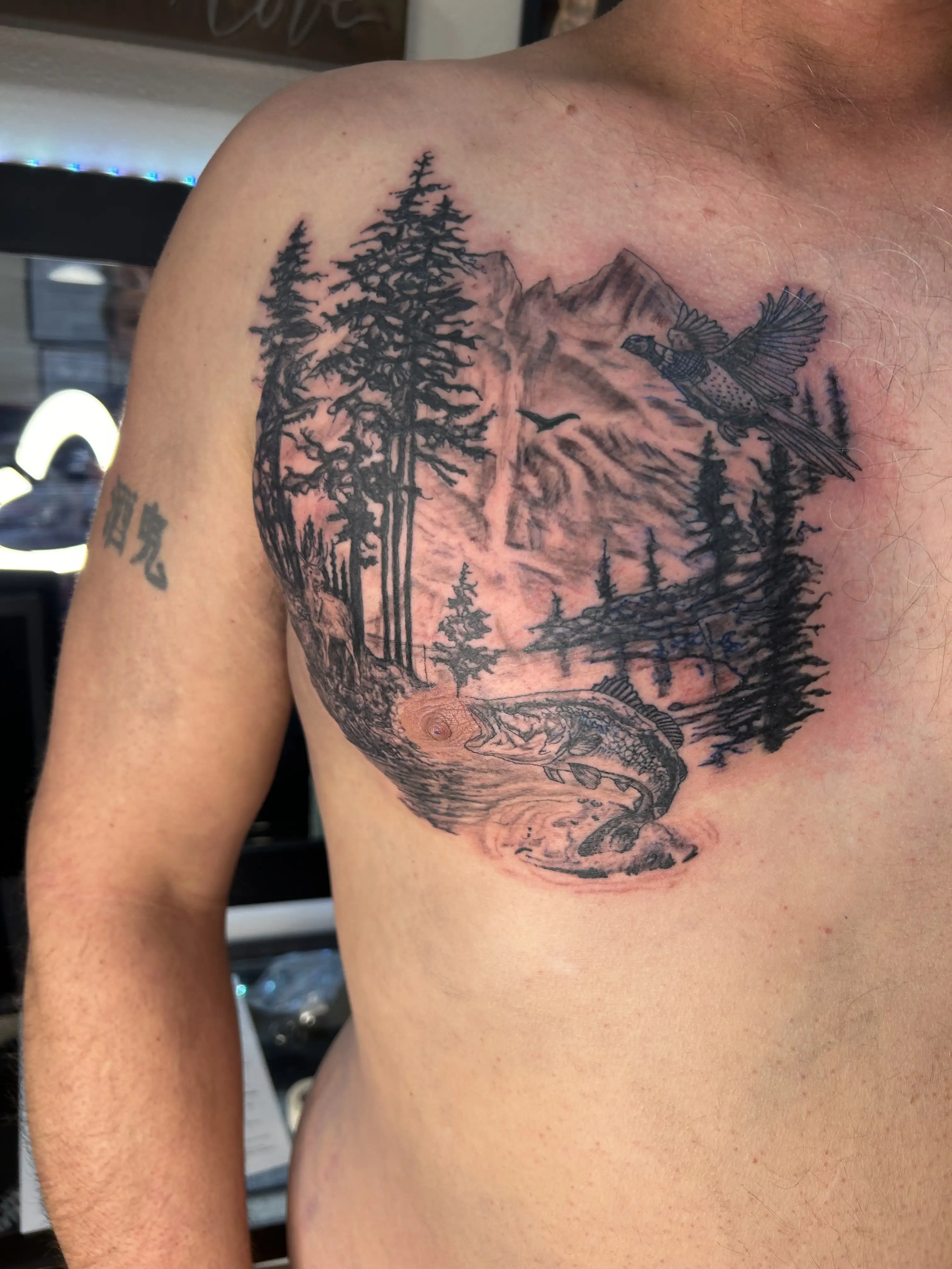BL-ink Tattoo Colorado Springs chest piece: black-grey mountain landscape with tall pines, a flying pheasant, and a trout leaping from the lake below.